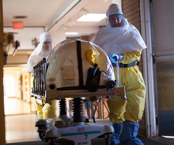 Two people in biocontainment suits rolling a device out a pair of doors