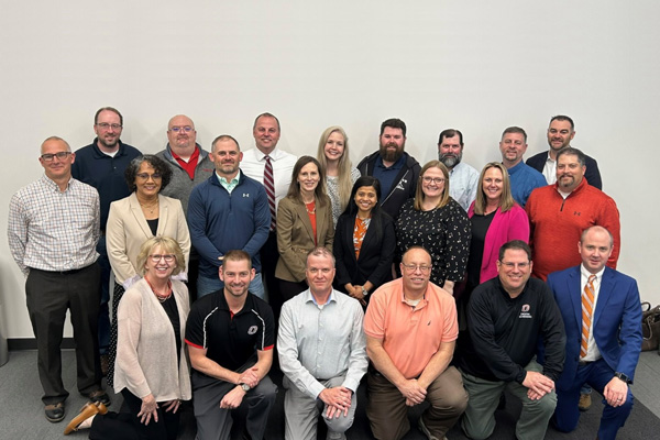 Large group of NU ITS staff posed in rows indoors against a plain wall, wearing business‑casual attire, with some seated in front and others standing behind.