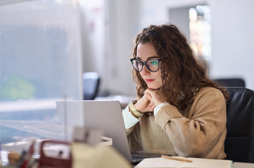 A person with shoulder‑length curly hair sits at a desk, looking at a laptop screen. The individual is wearing a light-colored sweater and is seated in an office environment with a protective screen and office supplies visible nearby.