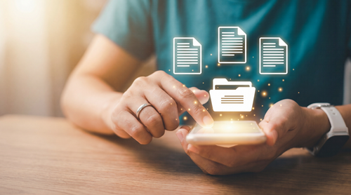 Close-up image of a person holding a smartphone in one hand while tapping the screen with the other hand. Above the glowing phone screen, digital-style icons appear, including several document icons and one folder icon, all floating as if being selected or accessed. The background is softly blurred, with warm lighting coming from the left side of the image.