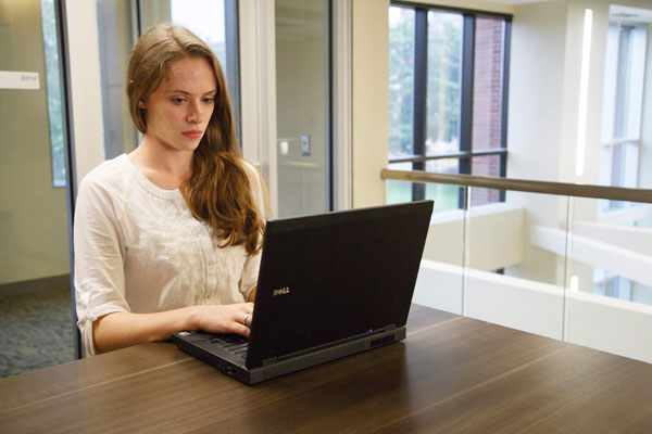 A person sitting at a wooden table in a bright indoor space, using an open Dell laptop. Large windows and glass panels surround the area, showing parts of a modern building interior with natural light coming in.
